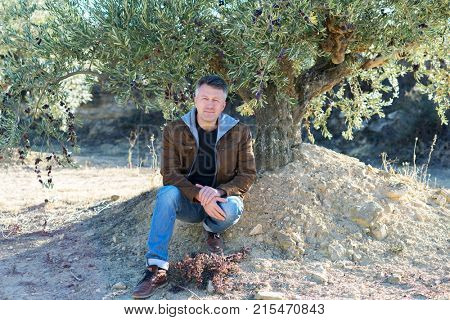 Handsome man posing in olive trees garden. Male portrain over mediterranean olive field ready for harvest. Confident mature man in spanish olive's grove with ripe fresh olives.