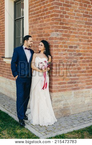 Handsome Male Bridegroom Looks With Great Love At His Bride. Happy Newlyweds Stand Near Brick Wall O
