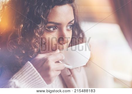 Let me think. Beautiful African woman with curly hair holding cup near mouth while drinking tea being in cafe