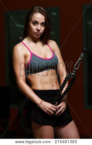 Charming sportive young woman in sportswear posing and looking at camera holding the handle of fitness machine in the gym