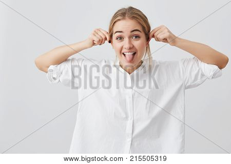 Playful funny pretty girl with gloomy expression demonstaring tongue, pulling herself at ears, mocking at camera. Playful beautiful european female with fair hair posing against gray background. Positive emotions concept.