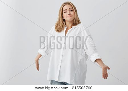 Blonde confused caucasian female in white shirt shrugging shoulders, stretching arms forward, staring at camera with questionable and clueless look, saying I don't know. People and emotions concept.