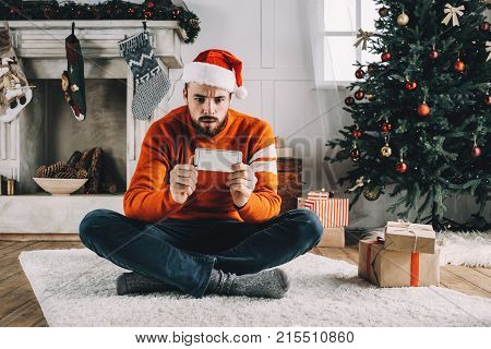 Nice and bearded young man is sitting on the carpet with his legs crossed and looking to the phone. He is watching Christmas special of his favourite TV-series and it's so interesting He can't wait what will happen next.