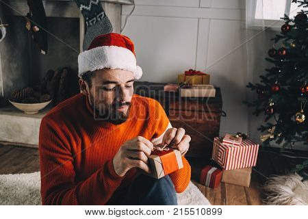 Nice and concentrated young man is sitting on the carpet on the floor and finishing packing presents for his friends and beloved. He is doing that very neatly and with some tengerness. Close up. Cut view