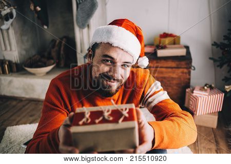 Close up of a bearded nice and cheerful guy sitting on the floor and presenting a gift that he packed by himself. Young man is happy to share some of his presents with somebody else. Cut view