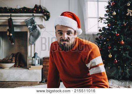 Funny picture of a bearded guy sitting on the floor in a white decorated room. Somehow he is not happy with something because his face shows such emotions as boring and unhappiness. He is looking straight to the camera. Close up