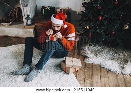 Cut view of a serious guy in red sweater sitting on the floor and packing a small gift for his friend. He is very concentrated on the work he is doing. He hopes his friend will like the present very much