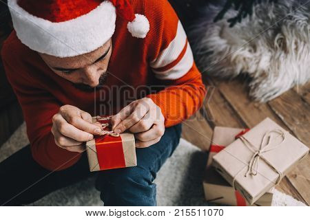 Close up of a young man in red hat and red sweater sitting on the wood brown floor and preparing a gift for his friend. He has packed the other two presents and now they are lying on the floor. Guy is concentrated on the packing. Cut view