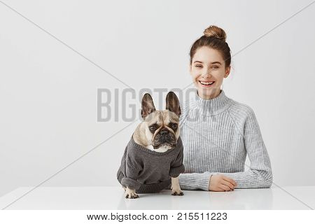 Portrait of smiling brunette woman with hair tied in topknot working at home office. Female freelancer sitting at the table in workshop in company of dog.  Friendship concept