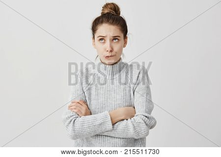 Young lady with hair in topknot standing with arms folded looking up. Capricious female model in grey dress biting lips thinking about her desire.  Daydream concept