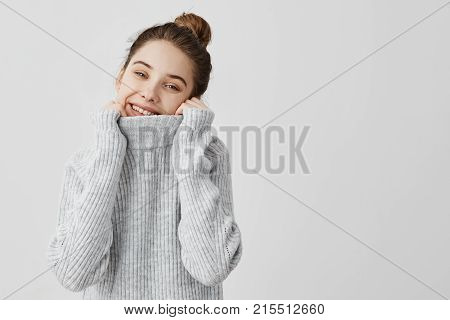 Beautiful teen girl hiding her neck in collar of woolen sweater with broad smile.  Female model posing in studio wearing cool warm outfit bending head to side. Joy and happiness concept