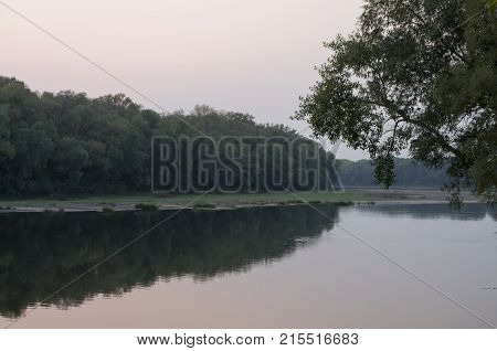 Wide river flowing across green forest. Fall. Evening. Reflections of trees in the calm water. Sundown