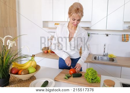 Attractive young woman cuts green cucumber for salad with sharp knife, takes red juicy tomato and several times throws up with smile on face, sniffs freshness of lettuce leaves and takes up shredding vegetables. Blonde at top of modern light kitchen with 