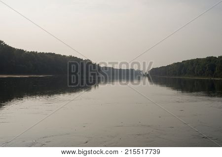 Wide river flowing across green forest. Fall. Evening. Reflections of trees in the calm water. Sundown