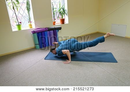 Young brunette of American origin sits on yoga mat and performs longitudinal twine. Short-haired sportswoman dressed in blue sports pants and blue t-shirt. Room light and spacious, under wall multi-colored yoga mats. Concept of to maintain body in good ph