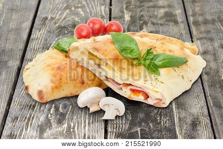Half and whole of the baked calzone - closed type of pizza that is folded in half decorated with basil twigs raw mushrooms and cherry tomatoes on an old rustic wooden surface