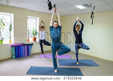 Two women and men of American appearance engaged in yoga, people sit on floor on fitness rugs and perform stretching exercises. Room light and spacious, walls of pleasant light color, on windowsills pots with flowers. Concept of maintain body in good phys