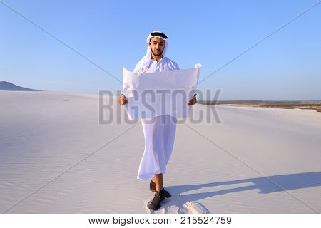 Beautiful Emirate Sheikh businessman studying project of future construction of complex, standing in middle of bottomless desert with white clean sand on clear warm day against blue sky. Swarthy Muslim with short dark hair dressed in kandura, long, spacio