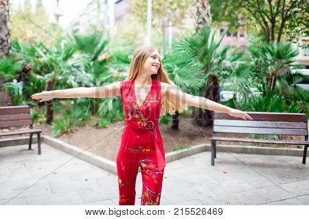 Carefree young lady dancing in summer park enjoying good weather. Happy girl in red jumpsuit spinning around outstretching arms. Slow life concept