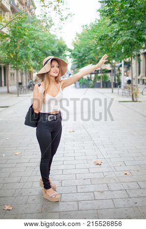 Pretty lady waving hand to catch cab while standing on street. Positive modern woman waiting for taxi. City life concept