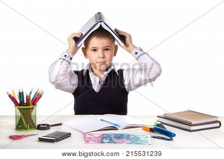 Smart serious excellent pupil at the desk with open book on the head on the white background.
