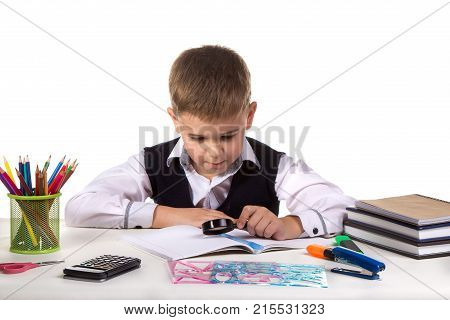 Smart serious pupil at the desk exploring with magnifier on the clean white background.