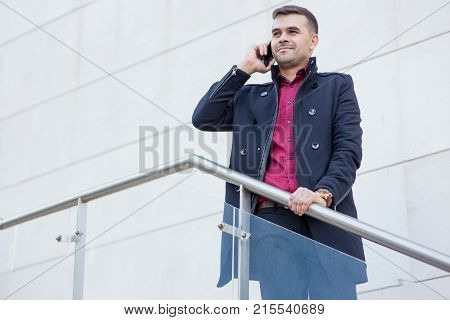 Portrait of confident young Caucasian businessman wearing coat standing on staircase and talking on mobile phone outdoors. Business and communication concept