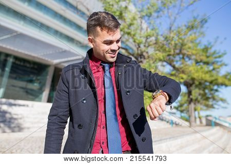 Portrait of happy young Caucasian businessman or student looking at watch and smiling outdoors. Education, business, punctuality concept