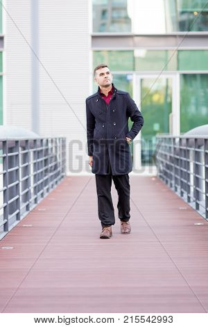 Portrait of serious young Caucasian businessman wearing coat walking along transition between offices outdoors. City and business life