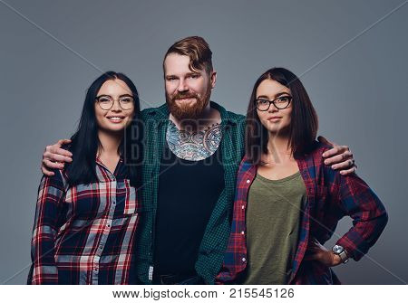 Bearded male with tattoos on his body and two brunette hipster women isolated over grey background.