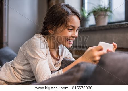 side view of smiling kid using smartphone while resting on sofa at home