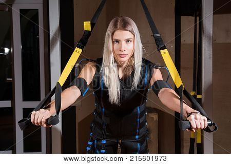 Blonde Girl In Electrical Muscular Stimulation Suit Doing Squat Exercise For Back And With Suspensio