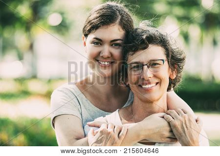 Portrait of cheerful Caucasian senior mother wearing glasses and her teenage daughter embracing, looking at camera and smiling outdoors