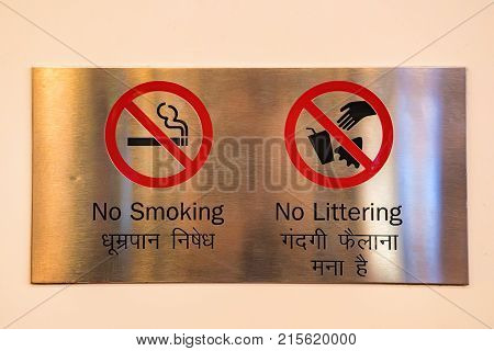 Close up two signs prohibiting smoking and littering on metal plate on white wall indoors