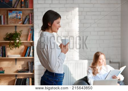 Young beautiful woman enjoying a cup of coffee smiling looking away her colleague working on a laptop on the background copyspace office cafe coffee shop workplace teamwork studying learn.