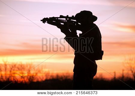 The soldier or hunter is aiming with cross-bow at sunset in nature silhouette.