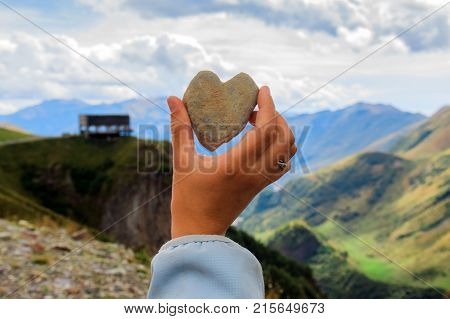 Arm holding the heart shaped stone in front of the wide view of high Caucasus Mountains beautiful landscape in Kazbegi , Georgia