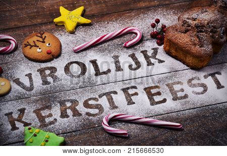 closeup of a table sprinkled with icing sugar or flour where you can read the text vrolijk kerstfeest, merry christmas in dutch, a rolling pin, some candy canes, a fruitcake and some christmas cookies