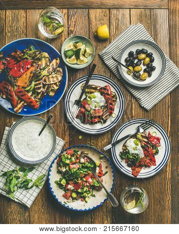 Flat-lay of healthy dinner table setting. Fresh salad, grilled vegetables with yogurt sauce, pickled olives, lemon water over wooden background, top view, square crop. Clean eating, vegetarian food
