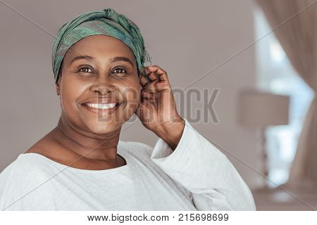 Close up of stylish mature African woman smiling while standing in her home putting on a traditional headscarf in a mirror