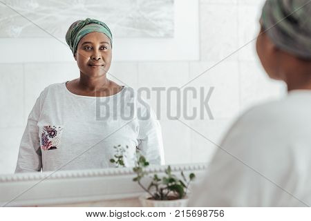 Stylishly dressed young African woman wearing a headscarf standing in her house looking at her reflection in a mirror