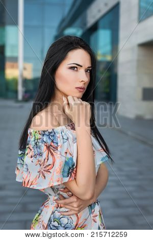Young thoughtfull beautiful brunette woman with long hair in the street wearing white dress with flower pattern on the background of skyscraper