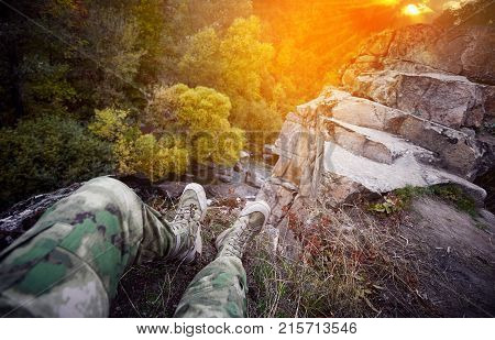 Traveler at a mountain river, legs over a cliff, outdoor clothing, deep stone canyon, ancient granite rocks, beautiful sunset, rocky river bank, mountain landscape, forest on the riverbank