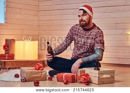 Redhead bearded male in Santa hat, drinks craft beer in a room with Christmas decoration.