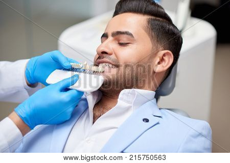 Shot of a handsome young man on a medical appointment at the dentist office professional dentist holding dentures comparing color and shade of teeth medicine healthcare whitening.