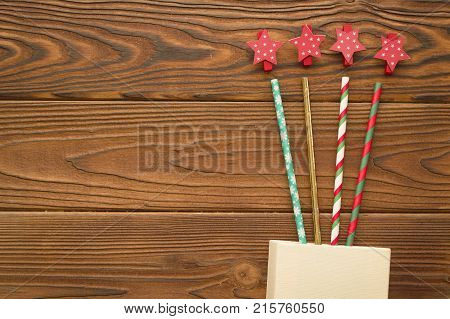 A beige box with straws with Christmas pattern and red star clips on a rustic wooden background. Christmas background. Copy space. Top view
