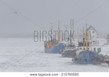 Ships of fishermen in the port on a frozen river in winter through a strong blizzard to the horizon