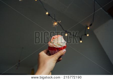 POV of female hand holding pink red velvet cupcake with sparkle candle burning concept burthday or romantic surprise for anniversary or date christmas lights