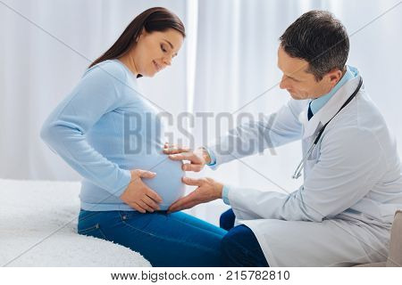 Let me see. Competent medical worker examining baby bump and bowing head while sitting opposite his patient