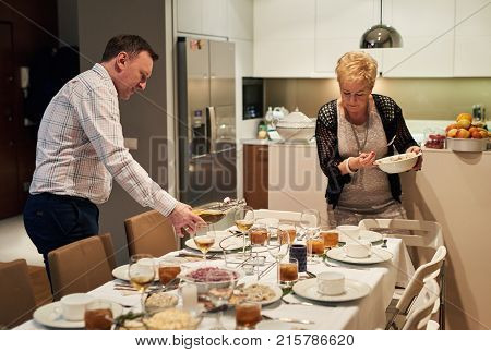 Happy family standing in a dining room setting a dining table and dishing out food and pouring drinks for christmas dinner at home